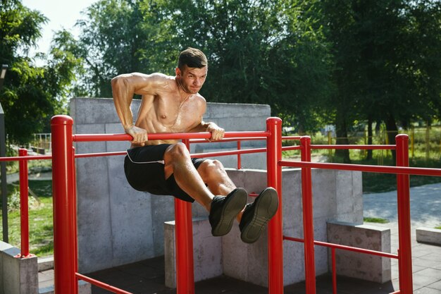 Hombre caucásico sin camisa muscular joven que hace dominadas en la barra horizontal en el patio en un día soleado de verano. Entrenando la parte superior del cuerpo al aire libre. Concepto de deporte, entrenamiento, estilo de vida saludable, bienestar.