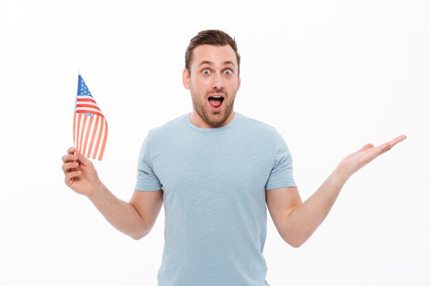 Hombre caucásico con cerdas sosteniendo pequeña bandera americana y levantando la mano en sorpresa