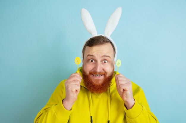 Hombre caucásico con decoración tradicional como un conejito de Pascua con ropa casual brillante sobre fondo azul de estudio. Felices saludos de pascua. Concepto de emociones humanas, expresión facial, vacaciones. Copyspace.