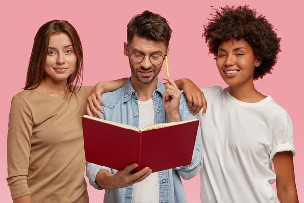 Un hombre caucásico desconcertado sostiene un libro de texto y un bolígrafo, intenta concentrarse en la tarea de escribir, dos mujeres de raza mixta se interponen entre él, tratan de ayudar, trabajan en equipo, modelan en la pared rosa. Concepto de diversidad