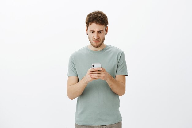 Hombre caucásico disgustado frustrado, sosteniendo un teléfono inteligente y mirando disgustado a la pantalla, haciendo muecas por las emociones negativas, no le gusta el nuevo artículo que leyó en el dispositivo