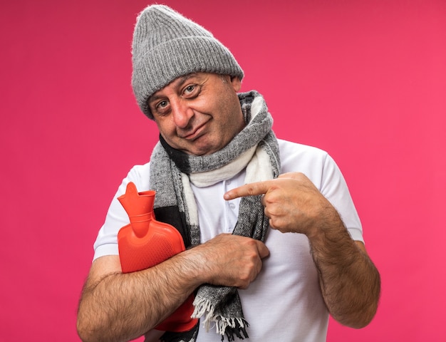 Hombre caucásico enfermo adulto complacido con bufanda alrededor del cuello con sombrero de invierno sosteniendo y apuntando a la botella de agua caliente aislada en la pared rosa con espacio de copia