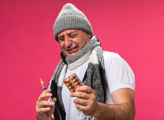 Hombre caucásico enfermo adulto disgustado con bufanda alrededor del cuello con sombrero de invierno se encuentra con los ojos cerrados sosteniendo una jeringa y un blister de medicina aislado en una pared rosa con espacio de copia