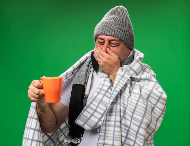 Hombre caucásico enfermo adulto disgustado con bufanda alrededor del cuello con sombrero de invierno envuelto en cuadros cerrando la nariz con la mano y mirando la taza aislada en la pared verde con espacio de copia