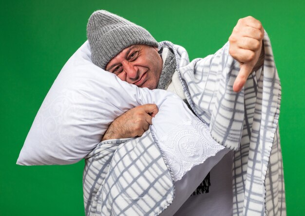 Hombre caucásico enfermo adulto disgustado con bufanda alrededor del cuello con sombrero de invierno envuelto en cuadros pulgares hacia abajo sosteniendo y poniendo la cabeza en la almohada aislada en la pared verde con espacio de copia