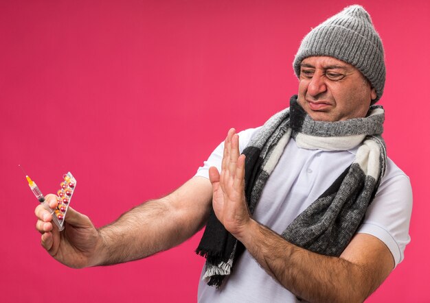 Hombre caucásico enfermo adulto disgustado con bufanda alrededor del cuello con sombrero de invierno sosteniendo y mirando el blister de jeringa y medicamento aislado en la pared rosa con espacio de copia