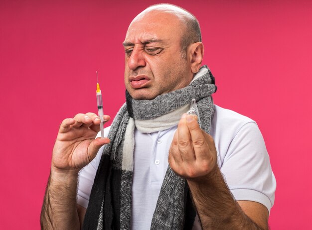 Hombre caucásico enfermo adulto disgustado con bufanda alrededor del cuello sosteniendo la ampolla y mirando la jeringa aislada en la pared rosa con espacio de copia