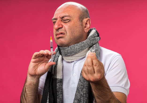 Hombre caucásico enfermo adulto disgustado con bufanda alrededor del cuello sosteniendo la ampolla y oler la jeringa aislado en la pared rosa con espacio de copia