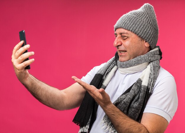 Hombre caucásico enfermo adulto molesto con bufanda alrededor del cuello con sombrero de invierno sosteniendo y apuntando al teléfono aislado en la pared rosa con espacio de copia