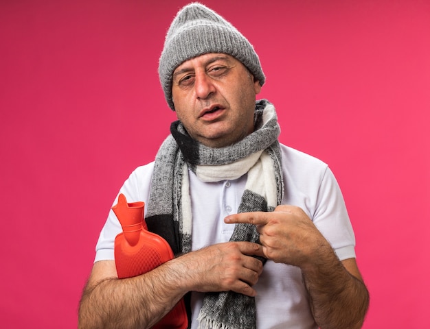 Foto gratuita hombre caucásico enfermo adulto molesto con bufanda alrededor del cuello con sombrero de invierno sosteniendo y apuntando a la botella de agua caliente aislada en la pared rosa con espacio de copia