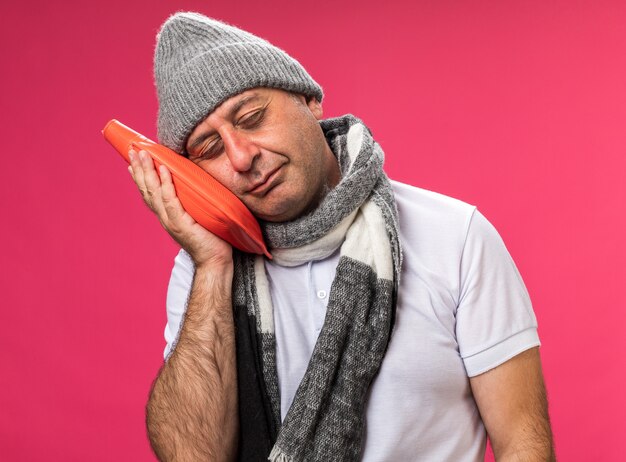 Hombre caucásico enfermo adulto soñoliento con bufanda alrededor del cuello con sombrero de invierno sosteniendo y poniendo la cabeza en la botella de agua caliente aislada en la pared rosa con espacio de copia