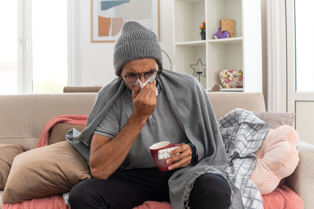 Hombre caucásico enfermo joven disgustado con gafas ópticas con sombrero de invierno se limpia la nariz con un pañuelo y sostiene una taza con un blister de medicina sentado en el sofá de la sala