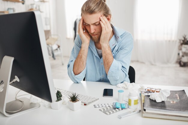 Hombre caucásico enfermo sentado en la oficina, apretando las sienes debido al dolor de cabeza, trabajando en la computadora, mirando la pantalla con expresión dolorosa en la cara, tratando de concentrarse, rodeado de medicamentos