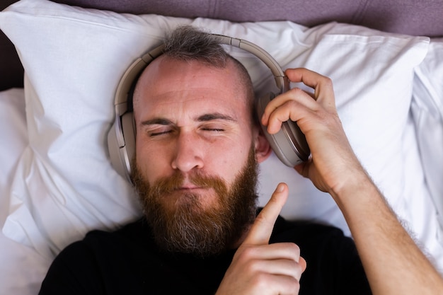 Hombre caucásico feliz en la cama con auriculares escuchando disfrutar de su música favorita, descansando solo, bailando.