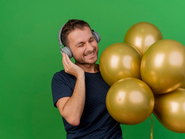 Hombre caucásico guapo alegre en auriculares sostiene globos de helio aislados sobre fondo verde con espacio de copia