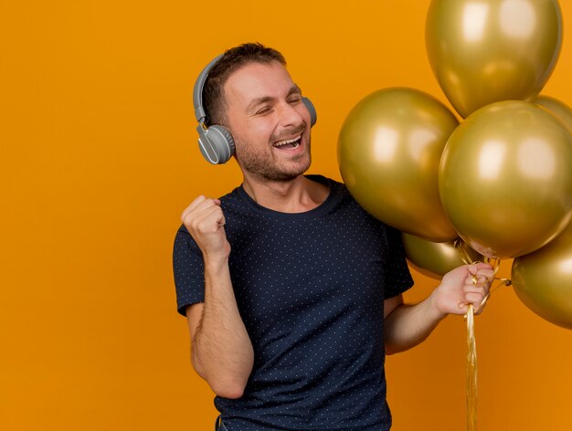 Hombre caucásico guapo alegre en auriculares sostiene globos de helio y mantiene el puño aislado sobre fondo naranja con espacio de copia