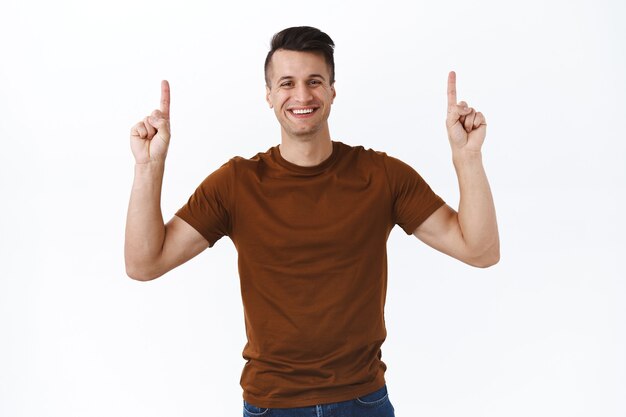 Hombre caucásico guapo alegre en camiseta marrón, consejos, haga clic en el enlace superior, señalando con el dedo hacia arriba y sonriendo