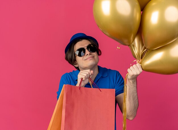 Hombre caucásico guapo complacido en gafas de sol con sombrero de fiesta azul sostiene globos de helio y bolsas de papel