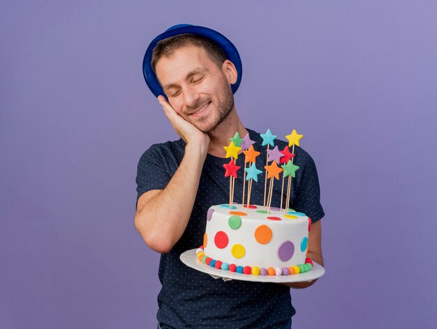 Hombre caucásico guapo complacido con sombrero azul pone la mano en la cara y sostiene el pastel de cumpleaños aislado sobre fondo púrpura con espacio de copia