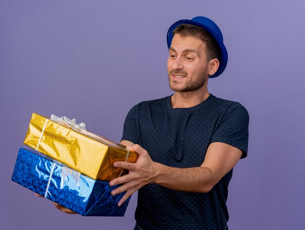 Hombre caucásico guapo complacido con sombrero azul sostiene y mira cajas de regalo aisladas sobre fondo púrpura con espacio de copia