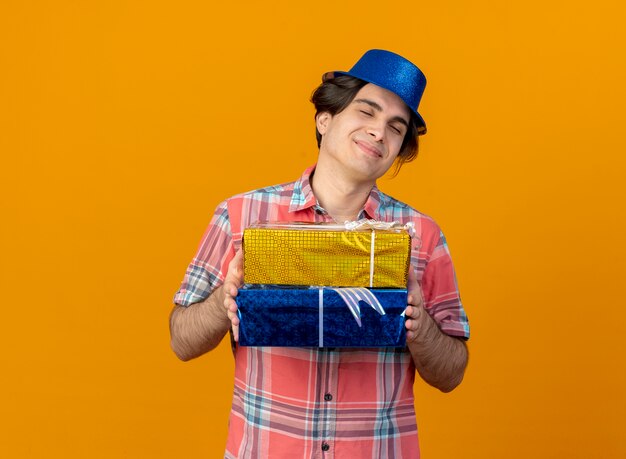 Hombre caucásico guapo complacido vestido con gorro de fiesta azul tiene cajas de regalo