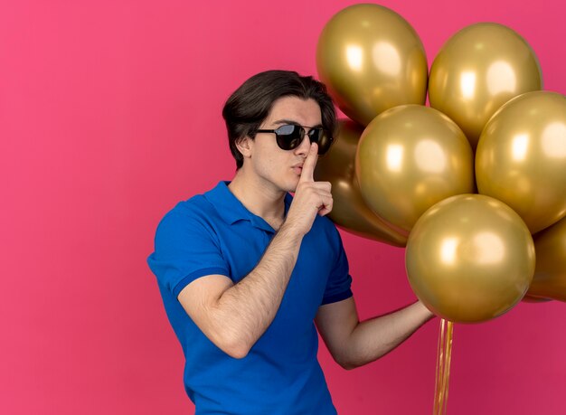 Hombre caucásico guapo confiado en gafas de sol sostiene globos de helio y gestos signo de silencio