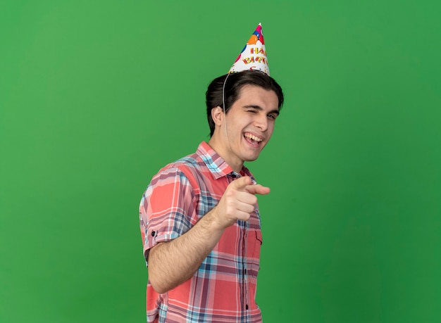 Foto gratuita hombre caucásico guapo confiado con gorro de cumpleaños parpadea y apunta a la cámara