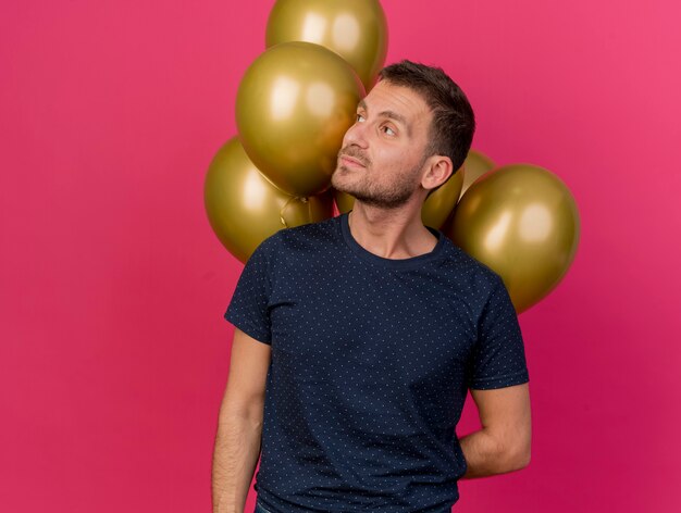 Hombre caucásico guapo confiado sostiene globos de helio detrás de la espalda mirando al lado aislado sobre fondo rosa con espacio de copia
