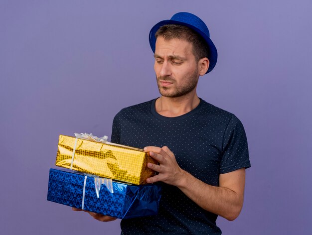 Hombre caucásico guapo disgustado con sombrero azul sostiene y mira cajas de regalo aisladas sobre fondo púrpura con espacio de copia