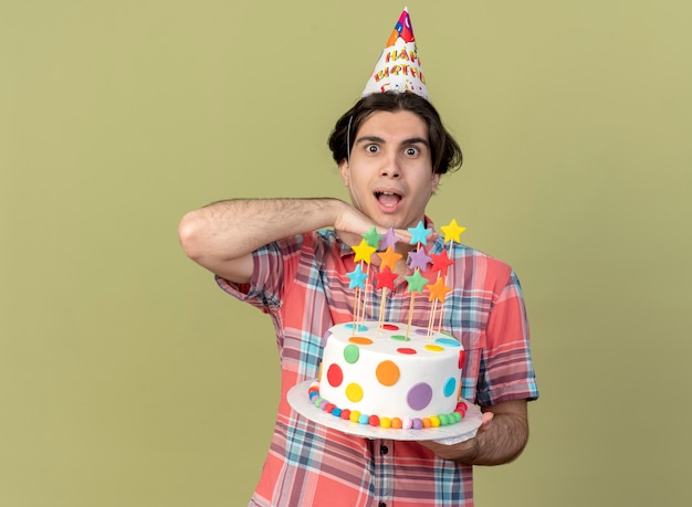 Hombre caucásico guapo emocionado con gorro de cumpleaños pone la mano en la barbilla y sostiene el pastel de cumpleaños