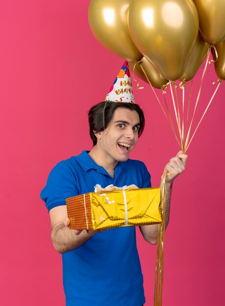 Hombre caucásico guapo emocionado con gorro de cumpleaños sostiene globos de helio y caja de regalo