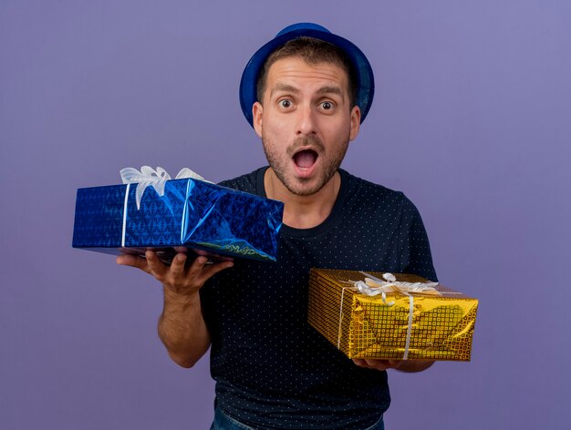 Hombre caucásico guapo emocionado con sombrero azul tiene cajas de regalo aisladas sobre fondo púrpura con espacio de copia