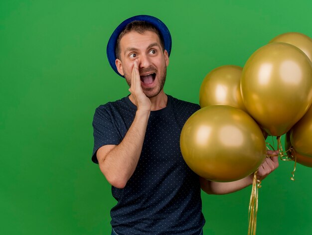 Hombre caucásico guapo emocionado con sombrero de fiesta azul sostiene globos de helio y mira al lado llamando a alguien aislado sobre fondo verde con espacio de copia