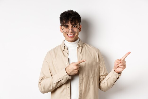 Hombre caucásico guapo con gafas sonriendo, señalando con el dedo en la esquina superior derecha, mostrando el logo sobre fondo blanco.