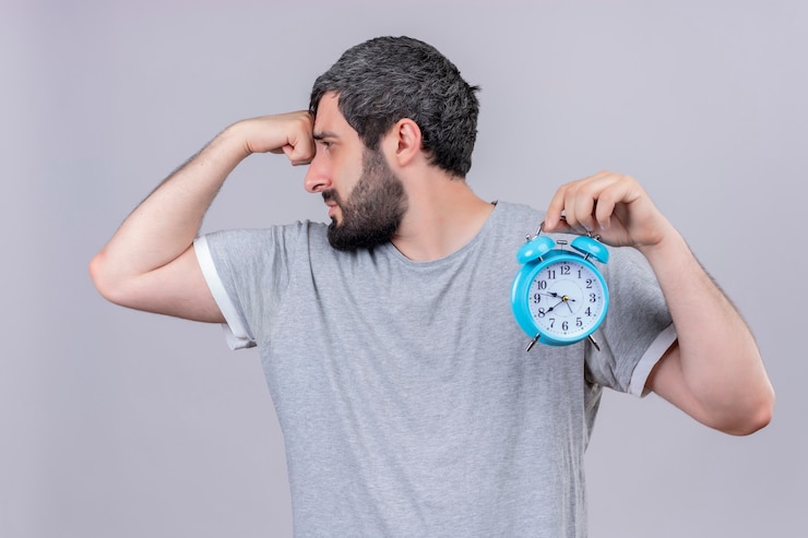 Foto gratuito hombre caucásico guapo joven disgustado sosteniendo reloj despertador mirando a un lado y poniendo la mano en la frente aislado sobre fondo blanco