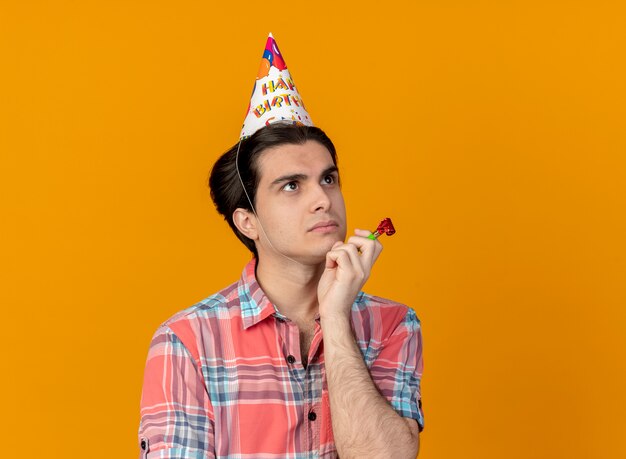 Hombre caucásico guapo pensativo con gorro de cumpleaños pone la mano en la barbilla sosteniendo un silbato de fiesta mirando al lado