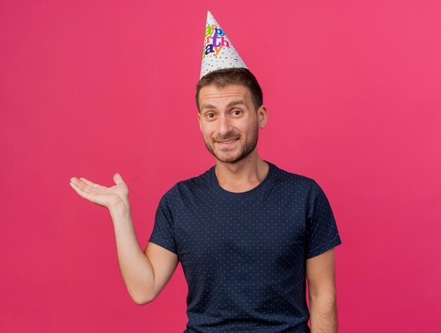 Hombre caucásico guapo satisfecho con gorro de cumpleaños sostiene la mano abierta aislada sobre fondo rosa con espacio de copia