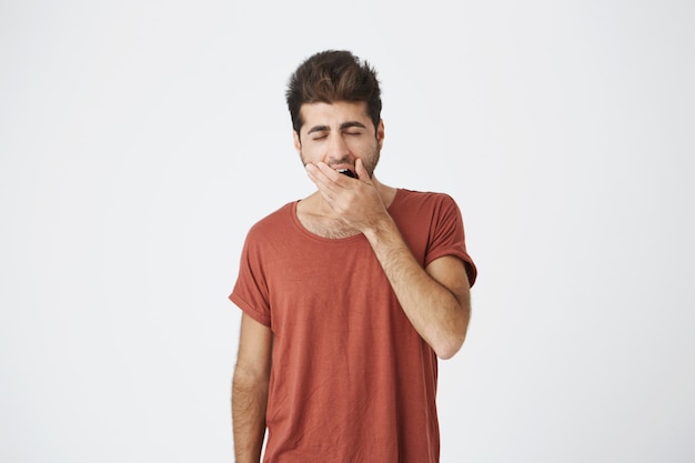 Hombre caucásico hermoso soñoliento que lleva la camiseta roja que cubre la boca mientras que bosteza, después de la conferencia aburrida en la universidad. Concepto de personas y emociones