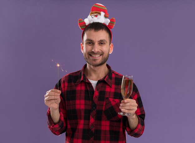 Hombre caucásico joven alegre con diadema de santa claus con bengala de vacaciones y copa de champán mirando a cámara aislada sobre fondo púrpura