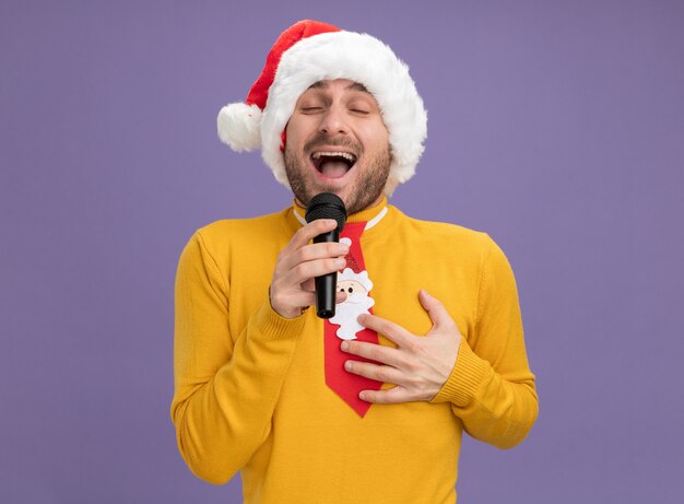 Hombre caucásico joven alegre con sombrero de navidad y corbata sosteniendo micrófono cantando con los ojos cerrados manteniendo la mano en el pecho aislado sobre fondo púrpura