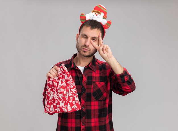 Hombre caucásico joven confiado con diadema navideña sosteniendo saco de navidad mirando a cámara mostrando el símbolo de signo v cerca del ojo guiñando un ojo aislado sobre fondo blanco con espacio de copia