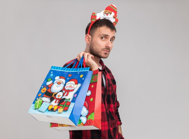Hombre caucásico joven confidente con diadema de Navidad de pie en vista de perfil sosteniendo bolsas de regalo de Navidad en el hombro mirando a cámara aislada sobre fondo blanco con espacio de copia