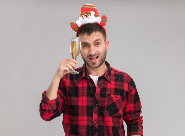 Hombre caucásico joven confidente con diadema de Navidad sosteniendo una copa de champán tocando la cabeza con ella mirando a cámara aislada sobre fondo blanco