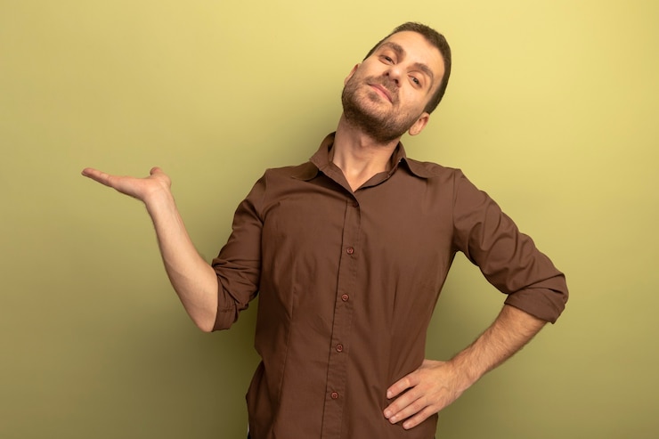 Foto gratuito hombre caucásico joven confidente que mira la cámara manteniendo la mano en la cintura que muestra la mano vacía aislada en fondo verde oliva