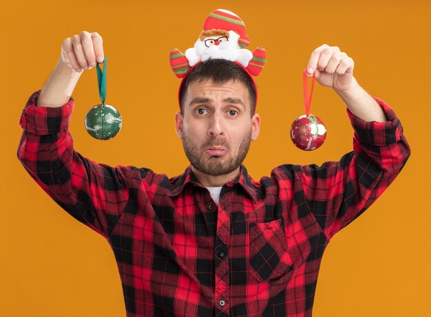 Foto gratuita hombre caucásico joven despistado con diadema de santa claus mirando a la cámara sosteniendo adornos navideños aislado sobre fondo naranja