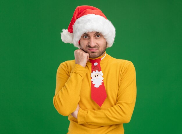 Hombre caucásico joven disgustado con sombrero de navidad y corbata poniendo la mano en la cara mirando a cámara aislada sobre fondo verde con espacio de copia