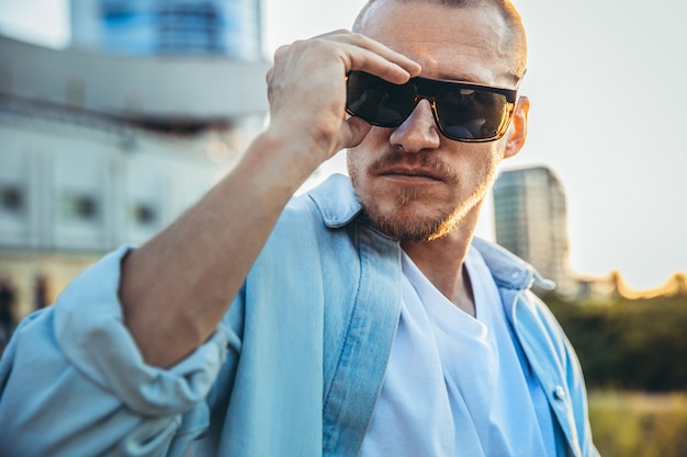 Hombre caucásico joven con gafas de sol, tiro moderno en efecto de grano de película y estilo vintage. Puesta de sol en las noches de verano. Calle de la ciudad. Tonos en verde azulado. Parece seguro y genial.