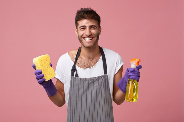 Hombre caucásico joven hermoso del servicio de limpieza listo para ordenar su apartamento hasta que esté tan limpio como la cera, equipado con detergente y esponja, mirando con una sonrisa feliz y alegre