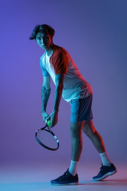 Hombre caucásico joven jugando tenis aislado sobre fondo de estudio púrpura azul en acción de neón y concepto de movimiento