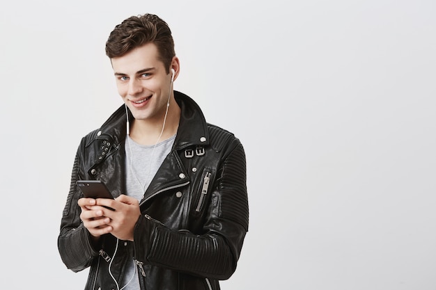 Hombre caucásico joven moderno con cabello oscuro en mensajes de chaqueta de cuero negro a través de las redes sociales, utilizando internet 3g en un dispositivo electrónico, mirando con sus ojos azules y una sonrisa atractiva.